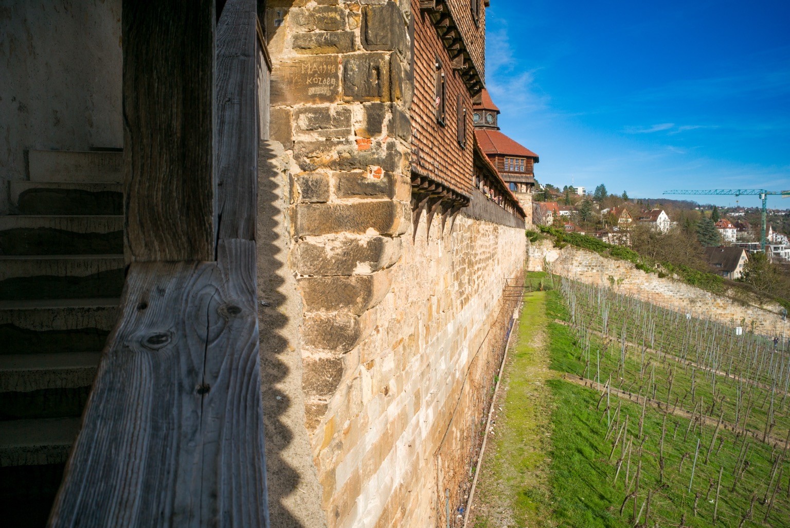 Ein dreigeteiltes Foto, das im linken Drittel Treppen der Burgstaffel, im mittleren Teil die Mauer der Esslinger Burg und im rechten Drittel den besagten Weinberg zeigt.