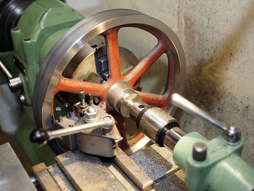 Das Foto zeigt ein Keilriemenrad, das auf einer Drehbank in ein Dreibackenfutter eingespannt und von einem Körner gegengehalten ist. Ein Drehstahl arbeitet an einer Passung.