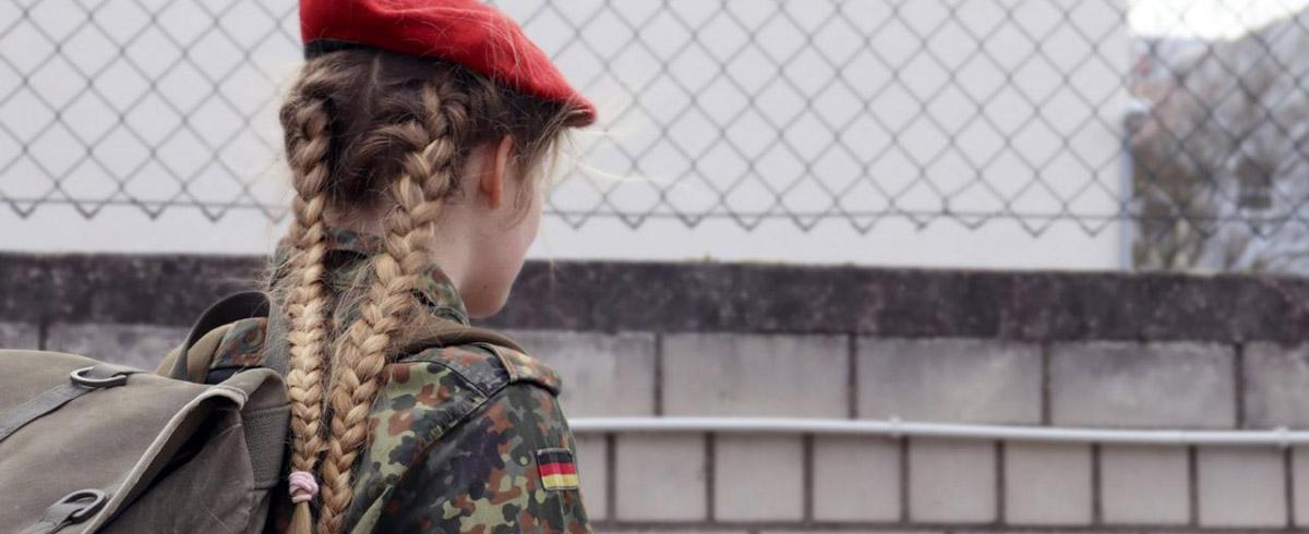 Das Foto zeigt eine Soldat*in von seitlich hinten rechts mit rotem Barett und Flecktarnbluse. Sie trägt einen Rucksack und blickt in Richtung einer Mauer, über der ein Maschendrahtzaun gesetzt ist. Ihre Haare sind zu zwei Zöpfen geflochten.