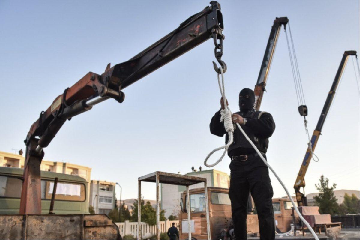 Eine öffentliche Hinrichtung auf dem Fayyam-Platz in Shiraz, 21. November 2018. (Students' News Agency)

Das Foto zeigt einen Vermummten, der auf einer LKW Ladefläche einen Galgenstrick hält, der an einem Kran des LKWs befestigt ist. Hinter ihm stehen weitere derartige LKWs.