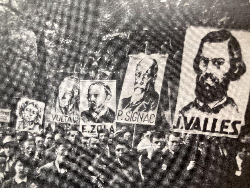 Das historische Foto zeigt die Menschenmassen vor dem Friedhof mit den Portraits verschiedener Persönlichkeiten der französischen Arbeiter- und Volksbewegung wie Voltaire, Emile Zola, Jules Vallès, Paul Signac