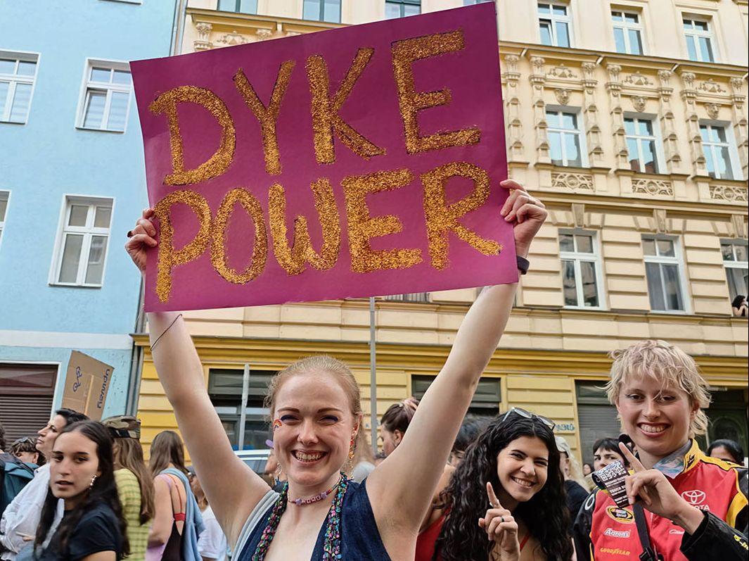 Das Foto zeigt einige Teilnehmer*innen des Marsches, die in die Kamera lächeln. Eine hält eine Papptafel mit dem mit Glitzergold geschriebenen Text "Dyke Power" nach oben.